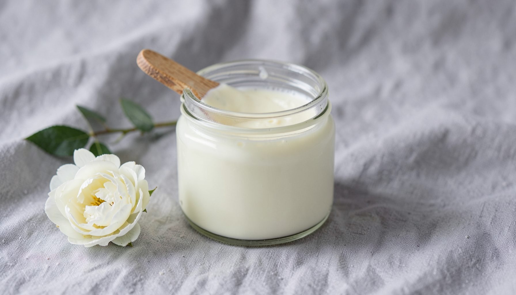 A glass jar of cream cleanser with a wooden spatula on a soft grey linen backdrop, accented by a single white peony