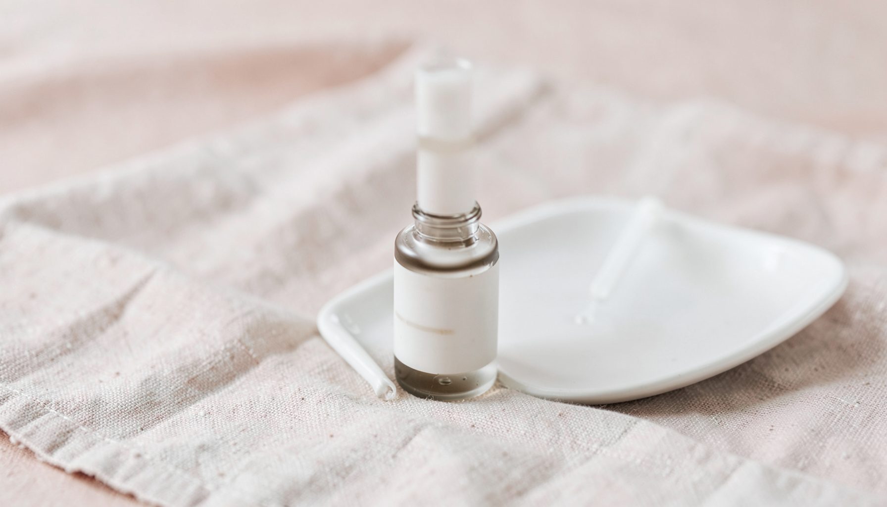 A close-up of a serum bottle with a dropper beside a white ceramic dish on a linen cloth, in soft neutral and blush tones.