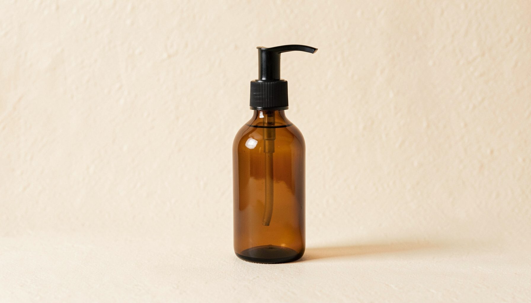 A minimalist amber glass bottle with a black pump on a textured cream backdrop, warm earthy tones.